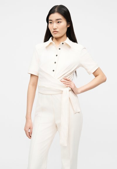 Γυναίκα με μακριά σκούρα μαλλιά που φοράει μια εκρού ολόσωμη φόρμα με κοντά μανίκια, κουμπιά και υφασμάτινη ζώνη δεμένη στη μέση.