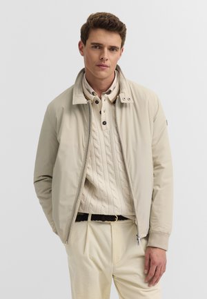 Junger Mann trägt eine beige Bomberjacke über einem cremefarbenen Pullover mit Zopfmuster und hellen Hosen, steht vor einem einfarbigen Hintergrund.
