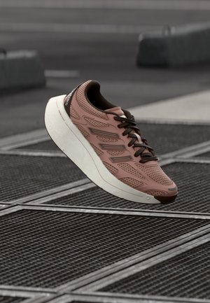 Chaussure de sport rose clair avec des lacets marron foncé et une semelle blanche flottant au-dessus d'une surface en grille métallique en extérieur.