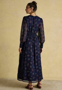 Tumes lilleline maksi kleit läbipaistevade pikkade varrukeidega, kogutud talje ja värvikirevad punaste ja siniste lillede mustrid. Looduslikud sandaalid täiendavad välimust.