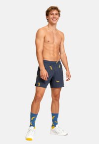 Marineblå svøm shorts med gule bananmønstre, elastisk talje, parret med blå sokker med bananmotiver og hvide sneakers.