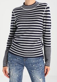 Navyblauer und grauer gestreifter Pullover mit langen Ärmeln und hohem Kragen, kombiniert mit verwaschenen blauen Jeans mit Perlenverzierungen.