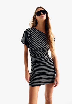 Frau mit großen schwarzen Sonnenbrillen und einem kurzen, einseitig schulterfreien schwarz-grau gestreiften Kleid, die vor einem schlichten weißen Hintergrund steht.