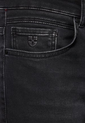 Zbliżenie na czarne dżinsy pokazujące małą przednią kieszeń z haftowanym krzyżem i szczegółowym ściegiem szwów.