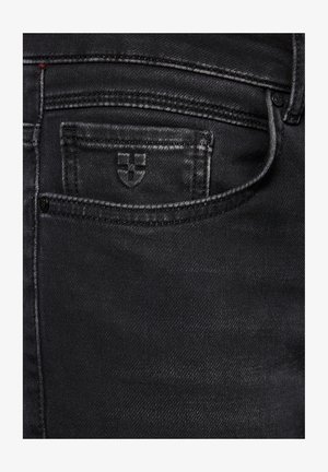 Zbliżenie na czarne dżinsy pokazujące małą przednią kieszeń z haftowanym krzyżem i szczegółowym ściegiem szwów.