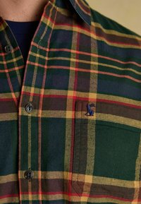 Četrdesmitdeviņu kreklu, kas ir raibā apdarē ar zaļiem, dzelteniem un sarkaniem toņiem, ar pogu aizdari, fakturētu audumu un mazu zilu izšūtu logotipu uz kabatas.