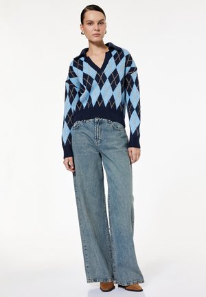 Femme portant un pull argyle bleu et marine avec col, un jean large bleu clair et des chaussures marron, debout devant un fond blanc.