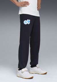 Marineblauwe trainingsbroek met een gladde textuur, voorzien van blauwe en witte logo's. Gecombineerd met witte sportschoenen met lichtbruine zolen en klittenbandsluitingen.