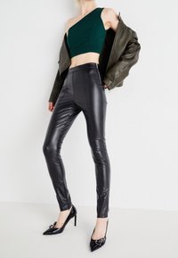 Kvinna som bär svarta läderleggings, grön axelbandslös crop top, olivgrön jacka som hänger över axlarna och svarta högklackade skor med spännen.