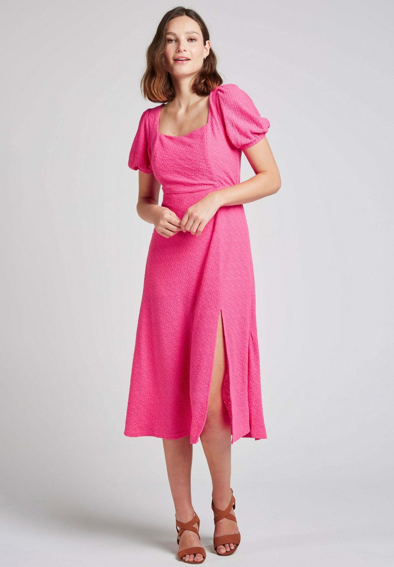 Robe midi texturée rose avec un décolleté carré, des manches bouffantes et une fente sur le côté. Modèle portant des sandales à lanières marron.