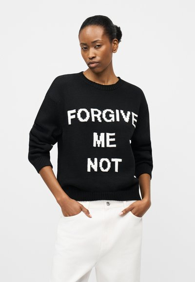 Γυναίκα που φοράει μαύρο πουλόβερ με λευκό κείμενο "ΜΗ ΜΟΥ ΣΥΓΧΩΡΕΙΣ" και λευκό παντελόνι, στέκεται με τα χέρια στις τσέπες μπροστά σε απλό φόντο.