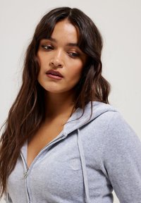 Felpa con zip di un grigio chiaro, dal texture morbido, con logo ricamato e tirazip in metallo. Include un cappuccio e cordini.