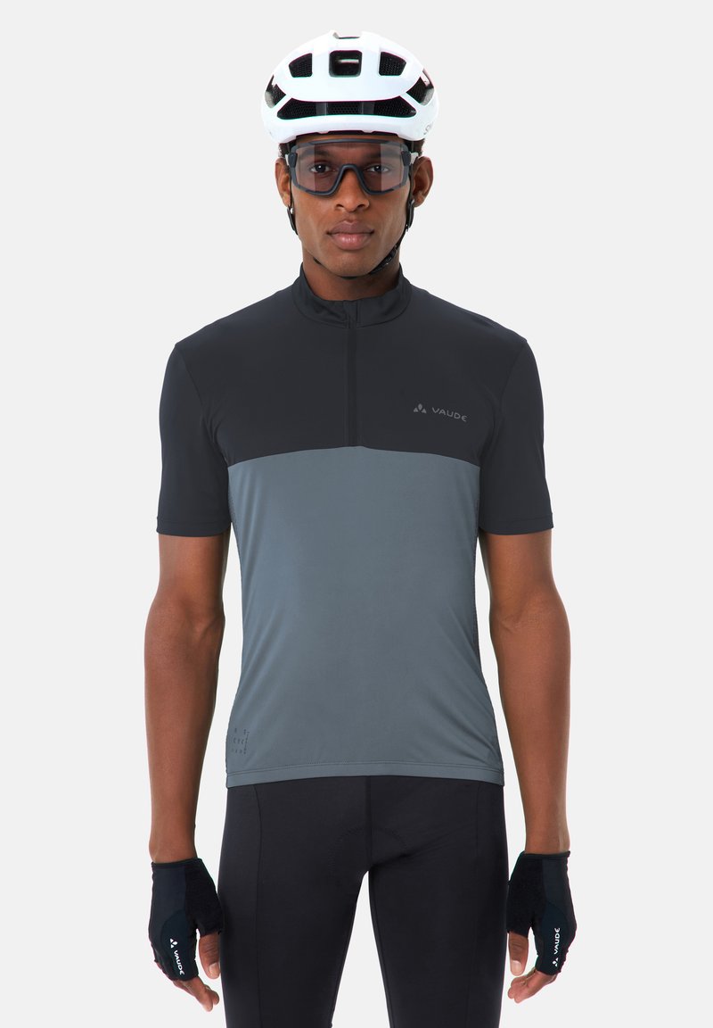 Fietser met witte helm, beschermende bril, zwart-grijs wielershirt, zwarte korte broek en zwarte vingervrije handschoenen, rechtopstaand.