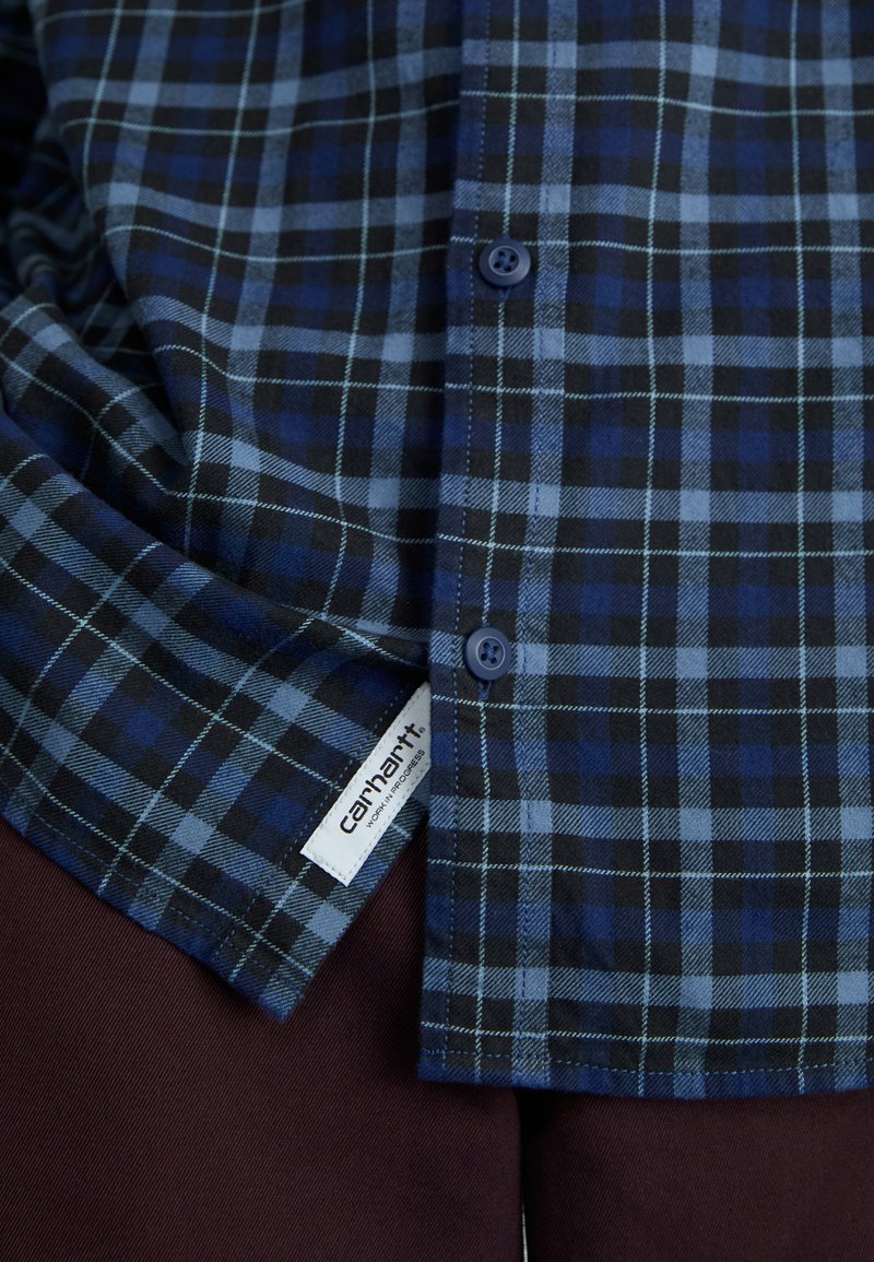 Blå och svart rutigt skjorta med knappar och en etikett som visar "Carhartt," kombinerad med mörk burgunderbyxor som har subtila texturer och mönster.