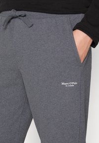 Nahaufnahme von grauen Sweatpants mit einem Kordelzugbund und einer Hand in der seitlichen Tasche, die ein weißes Marc O’Polo-Logo auf dem Oberschenkel zeigen.