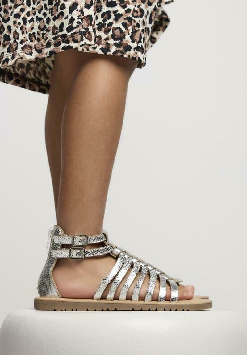Des sandales argentées à lanières avec une texture reptilienne, dotées de doubles brides de cheville et d'une semelle plate marron clair. Le design est ouvert à l'avant et respirant.