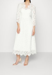 Valkoinen pitsinen midimekko, jossa on vartalonmyötäinen yläosa, kolmasosa hihat ja aaltoileva helma. Yhdistetty vaaleisiin sandaaleihin.