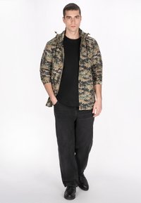 Kamuflagejakke med justerbar hætte i beige og mørkegrønne nuancer, parret med en sort t-shirt og løstsiddende sorte jeans.