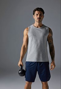 Raumeningas vyras su tatuiruotėmis, vilkintis pilką be rankovių marškinėlį ir mėlynus šortus, dešinėje rankoje laiko juodą kettlebellą prieš pilką foną.