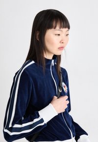 Giovane donna con capelli scuri lisci, che indossa una giacca blu navy con cerniera e strisce bianche e una piccola toppa ricamata.