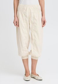 Pantalons crop beige clair avec taille smockée, caractérisés par des poignets élastiques, un tissu léger et une coupe décontractée. Associés à des ballerines à motifs.