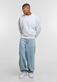 Hellgrauer, übergroßer Pullover mit gerippten Bündchen, kombiniert mit hellblauen, weiten Jeans und weißen Sneakers. Klare, eckige Brillengläser unterstreichen den Look.