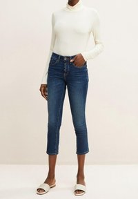 Vit långärmad topp och mörkblå cropped jeans med en figursydd design. Jeansen har knappstängning och horisontella sömmar ovanför knäna.
