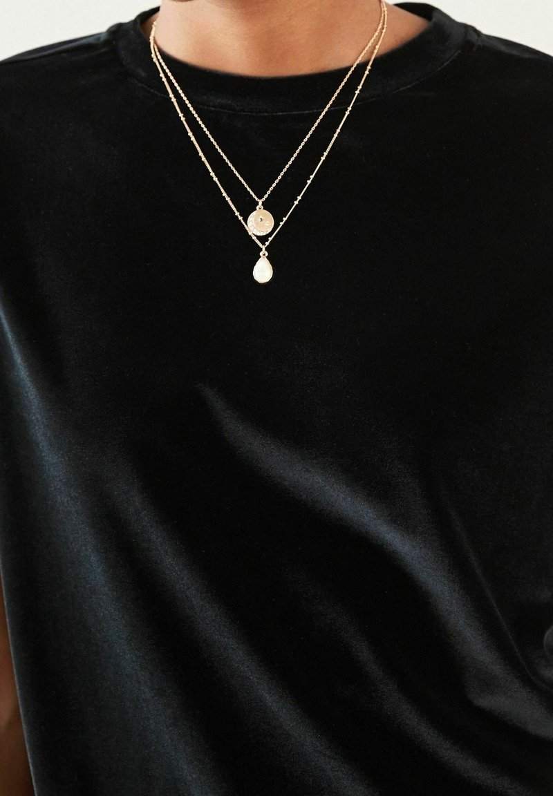 Dwuwarstwowy złoty naszyjnik z okrągłą medalionem i wisiorkiem w kształcie kropli noszony przez osobę ubraną w czarną aksamitną koszulę.