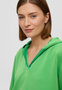 Zielona bluza z kapturem o dekolcie w kształcie litery V. Miękka tkanina o gładkiej teksturze. Posiada warstwowy kołnierz w nieco ciemniejszym odcieniu zieleni.