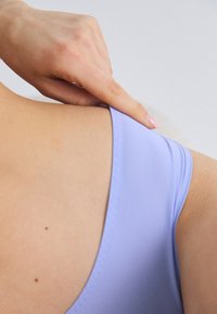 Costum de baie lila cu o textură netedă și mată. Prezintă o spate adânc și bretele subțiri. O mână trage ușor de material.