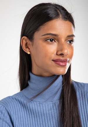 Modell trägt einen hellblauen, gerippten Rollkragenpullover. Goldene, kreisrunde Ohrringe betonen ihre Gesichtszüge. Glattes Haar fällt ihr über die Schultern.