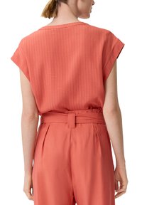 Korallfarbendes Top mit kurzen Ärmeln und strukturiertem Muster, kombiniert mit korallfarbenen, hoch geschnittenen Hosen mit einem Gürtel in der Taille und Faltendetails.