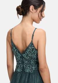 Turkusowa sukienka z głębokim dekoltem z tyłu, wyróżniająca się skomplikowanym haftem z koralików i cekinów. Lekka, teksturowana tkanina z delikatnym połyskiem.