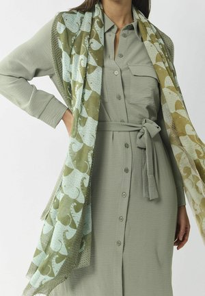 Femme portant une robe boutonnée vert clair avec une ceinture nouée à la taille et un grand foulard à motifs drapé sur ses épaules.