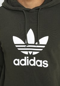Olivgrön huppartröja i bomull med en stor vit Adidas-logotyp med tre ränder ovanför och en treklöverdesign.