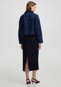 Chaqueta de mezclilla oscura recortada con un collar negro, combinada con un vestido midi negro ajustado que presenta una abertura lateral y tacones negros con tiras.