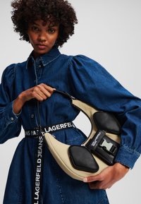Tan and black handbag with padded accents, curved shape, and a prominent buckle. Model wears a denim dress with puffed sleeves and logo belt.