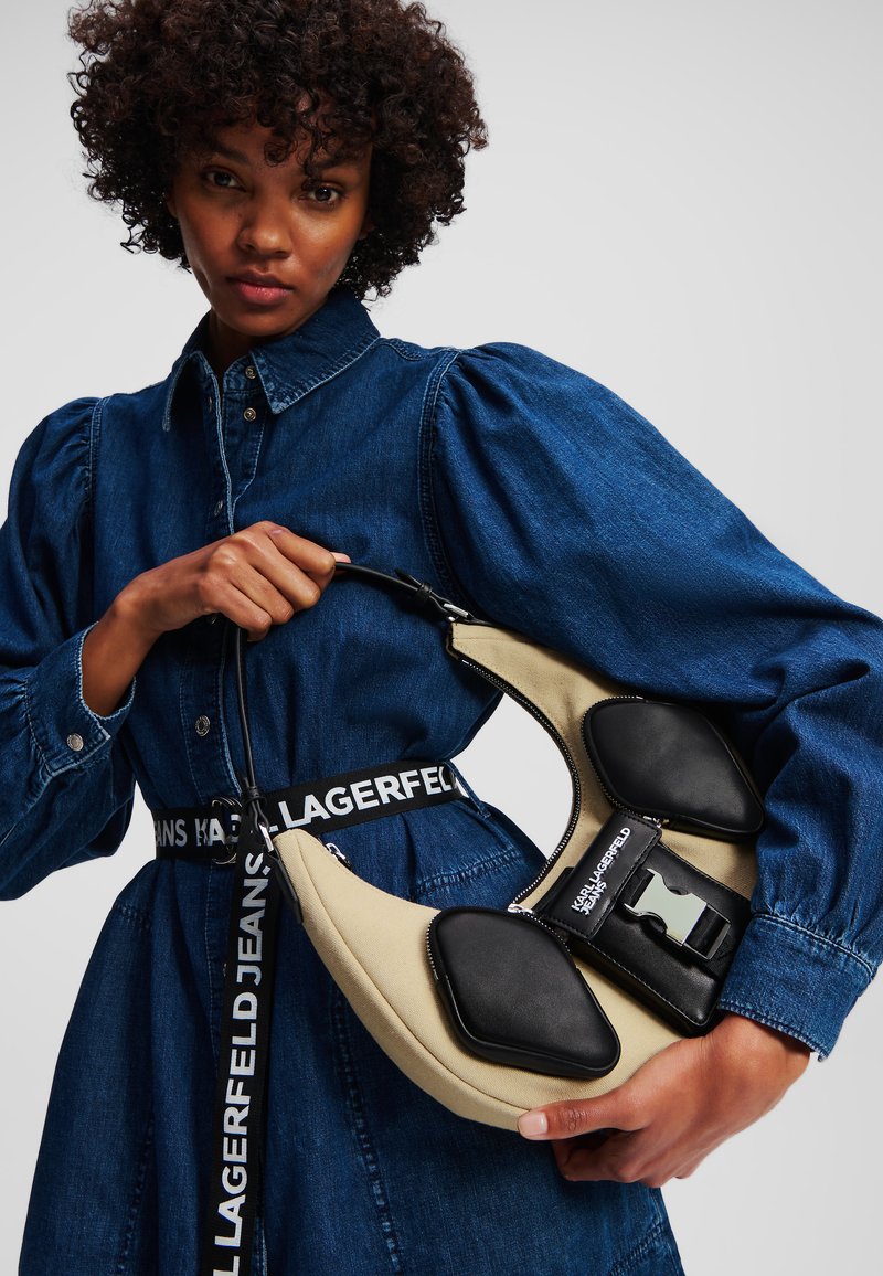 Tan and black handbag with padded accents, curved shape, and a prominent buckle. Model wears a denim dress with puffed sleeves and logo belt.