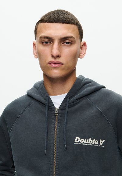 Junger Mann mit kurzen Haaren, der einen dunkelgrauen Kapuzenpullover mit Reißverschluss und dem Schriftzug "Double A" auf der Brust trägt, schaut direkt in die Kamera.