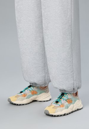 Hellgraue Jogginghose, an den Knöcheln aufgerollt, mit mehrfarbigen Turnschuhen, die beige, grüne, orangefarbene Akzente und türkisfarbene Schnürsenkel auf einem grauen Boden haben.