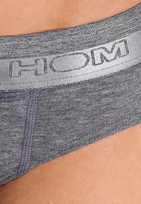 Biancheria intima da uomo grigia con un tessuto liscio e lavorato e una vita color argento con il nome del marchio "HOM" in lettere maiuscole.