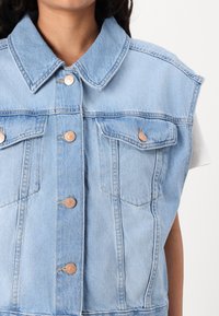Gilet in denim azzurro chiaro con colletto, due tasche sul petto e bottoni in bronzo. Presenta una vestibilità rilassata e una texture in denim.