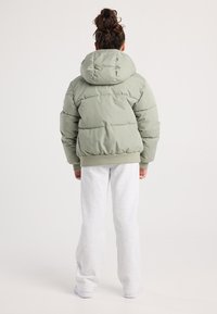 Persona con el pelo rizado recogido en un moño, vistiendo una chaqueta acolchada verde claro, pantalones de chándal blancos y zapatillas blancas, de pie de espaldas.