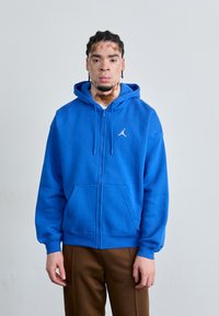 Blauwe zip-up hoodie met kangoeroezakken, geribbelde manchetten en een wit logo op de borst. Combinatie met bruine broek voor een complete look.