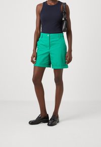 Grüne Baumwollshorts mit umgeschlagenen Säumen, kombiniert mit einem tiefblauen, ärmellosen Oberteil. Schwarze Slipper runden das Outfit ab. Einfaches, modernes Design.