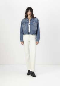 Casaco de denim cortado em azul médio com dois bolsos no peito, combinado com calças de perna reta em off-white e botas de tornozelo pretas.