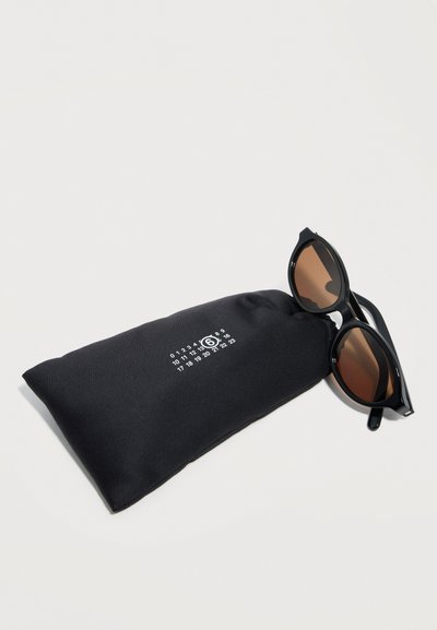 Black rounded sunglasses with brown lenses resting on a black soft fabric pouch with white printed numbers and a circled 6.