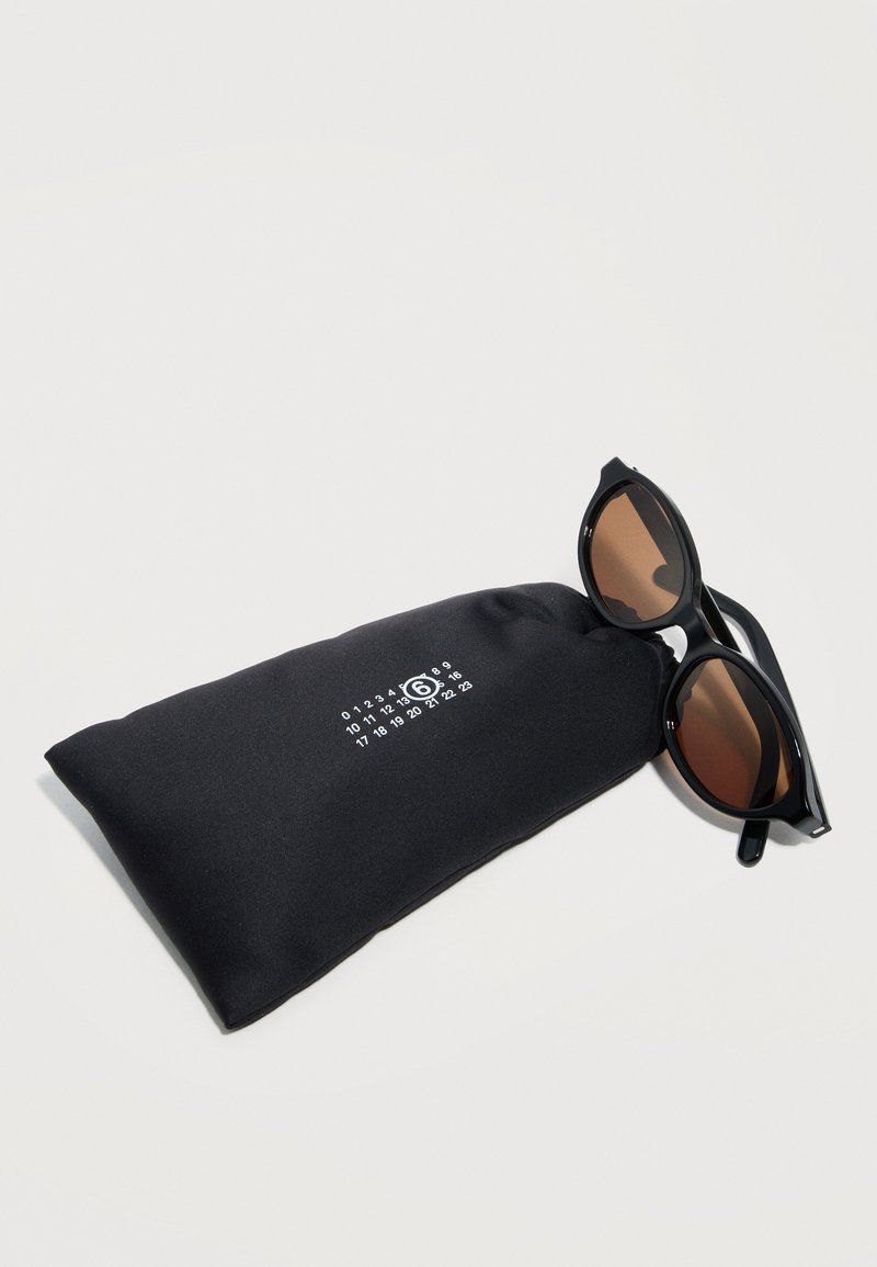 Black rounded sunglasses with brown lenses resting on a black soft fabric pouch with white printed numbers and a circled 6.