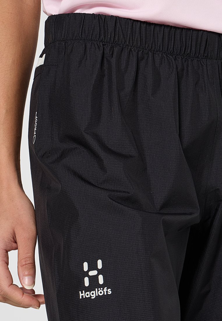 Mustat vedenpitävät housut, joissa on joustava vyötärö ja teksturoitu pinta. Sivulla on valkoinen logo ja heijastavia yksityiskohtia.