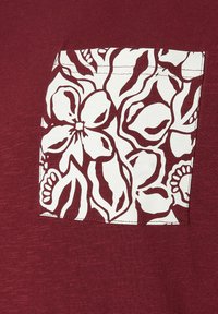 Tissu bordeaux avec une poche rectangulaire en patch au motif floral blanc cousue dessus, présentant de grandes formes de fleurs abstraites.